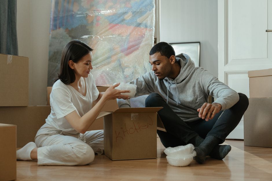 A man dressed in a grey hoodie is inspecting a cardboard box during a home relocation process inside a room with natural light coming through large windows covered partially by grey curtains. The room