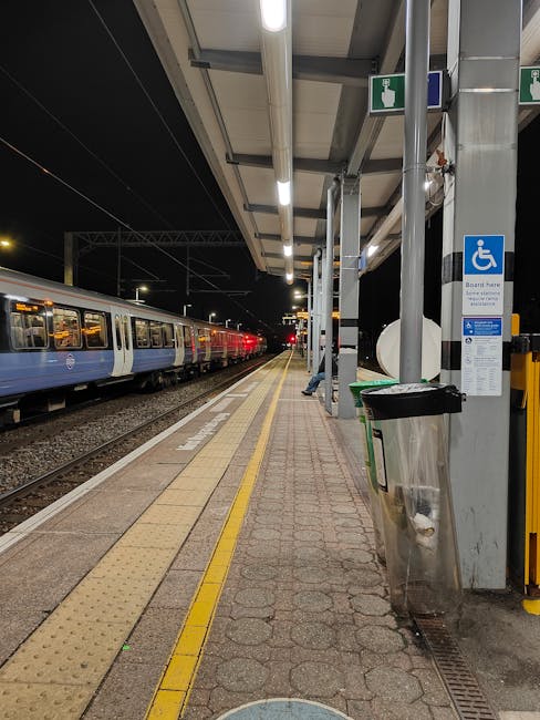 A view of Fulham Broadway station platform at night, illuminated by overhead lighting. The platform is paved with textured yellow tactile paving strips along the edge for safety, and a row of metal support columns extends along the length of the platform. Adjacent to the platform, there is a shelter with a roof supported by metal beams, providing cover for waiting passengers. A large blue and white accessible blue badge sign indicates wheelchair access, attached to one of the columns. Underneath the shelter, there is a black waste bin with a silver lid and a small green recycling container. To the left, a train with a blue and white exterior is stationary on the tracks, with its windows reflecting the interior lighting. The train's doors are closed, and the station environment suggests a typical overnight or early evening scene, potentially during a home relocation process involving moving furniture or boxes through the station. The setting is clean, organized, and captures the typical layout of a busy urban rail station. During a removals process, Movers Fulham would coordinate safe and efficient access for transport vehicles and moving equipment from this station area.