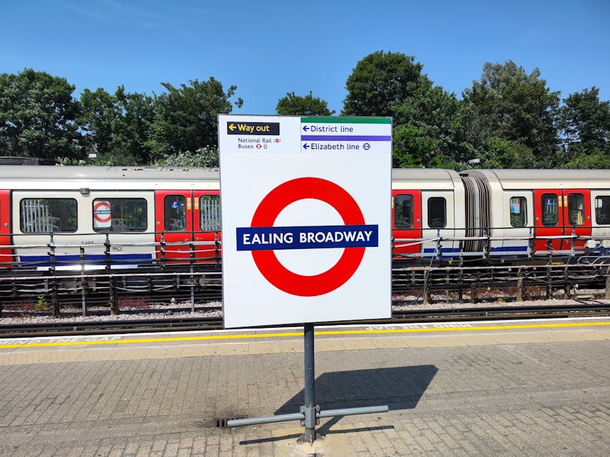 A large white sign with a red circle and blue horizontal bar displaying the station name 'Ealing Broadway' is positioned on a paved platform next to train tracks. Above the sign, smaller directional signs indicate the way out, district line, and Elizabeth line with arrows pointing in different directions. Behind the sign, a modern train with red, white, and blue livery is stationary on the track, with reflective windows and doors visible. The train is secured with a metal railing in front, and the scene is set outdoors on a bright, sunny day, with tall green trees and a clear blue sky in the background. The environment suggests a typical London Underground station area, suitable for disassembly or relocation considerations involved in house removals or moving services, as offered by Movers Fulham.