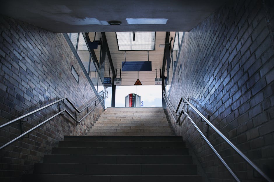 A view of Fulham Broadway station platform at night, illuminated by overhead lighting. The platform is paved with textured yellow tactile paving strips along the edge for safety, and a row of metal support columns extends along the length of the platform. Adjacent to the platform, there is a shelter with a roof supported by metal beams, providing cover for waiting passengers. A large blue and white accessible blue badge sign indicates wheelchair access, attached to one of the columns. Underneath the shelter, there is a black waste bin with a silver lid and a small green recycling container. To the left, a train with a blue and white exterior is stationary on the tracks, with its windows reflecting the interior lighting. The train's doors are closed, and the station environment suggests a typical overnight or early evening scene, potentially during a home relocation process involving moving furniture or boxes through the station. The setting is clean, organized, and captures the typical layout of a busy urban rail station. During a removals process, Movers Fulham would coordinate safe and efficient access for transport vehicles and moving equipment from this station area.