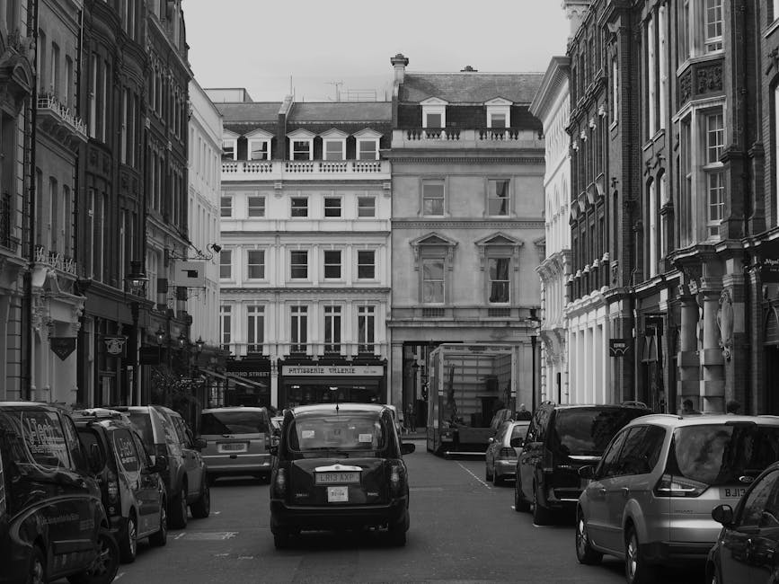 Black and white photograph depicting a narrow street in Fulham, London, with multiple parked cars along both sides of the road and a delivery truck positioned at the curb. The surrounding buildings are predominantly multi-storey residential properties with classic architectural details, including bay windows and decorative cornices. The scene captures an outdoor scene in daytime, with natural light illuminating the facades and vehicles. This environment highlights the logistical challenges of house removals in narrow access areas, such as King Street Fulham, where careful furniture transport and loading processes are essential. The presence of parked vehicles and the tight street width emphasize the need for specialized removals services that include packing, loading, and transportation of household items, often requiring precise maneuvering within restricted spaces. The image reflects the typical urban setting where professional movers like Movers Fulham operate, ensuring efficient and safe home relocation within such constrained environments.