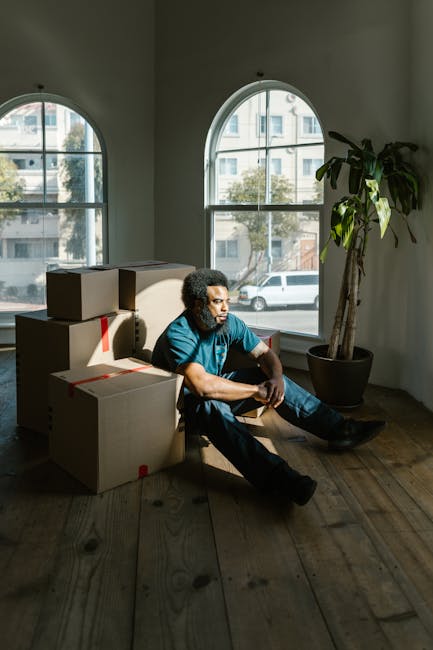 A man with curly dark hair and medium skin tone is sitting on the wooden floor of a room, surrounded by several cardboard boxes of varying sizes that are sealed with red packing tape, indicating they are packed for a home relocation. The boxes are stacked to his left, some leaning against the wall behind him. The room features two large, arched windows with white frames, allowing natural daylight to illuminate the space; through the windows, a street with parked cars and trees is visible. To the right of the man, there is a tall potted plant with broad, dark green leaves. The man is wearing a blue T-shirt and dark jeans, and appears to be resting during the packing and moving process, which is part of the furniture transport and logistics involved in a professional removal service. The overall setting reflects a moment in the packing and loading phase of a house removal, supported by the environment and contents that Movers Fulham might facilitate.