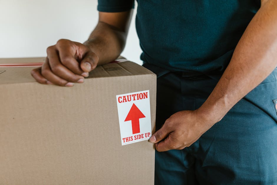 Close-up of a person’s hands securing a large cardboard box during a home relocation process, with the label 'Caution This Side Up' visible on the box. The individual is wearing a dark t-shirt and work pants, and the box is placed on a flat surface, possibly inside a property or at the entrance. The box appears to be part of a packing and moving activity, with the person carefully ensuring the correct orientation of the package, which may contain household items or delicate goods. Equipment such as packing tape and possibly a trolley or other moving tools are not visible but implied by the context. This scene is representative of professional furniture transport and loading procedures carried out by movers like Movers Fulham, supporting efficient and careful house removals in Fulham, SW6, and surrounding areas.