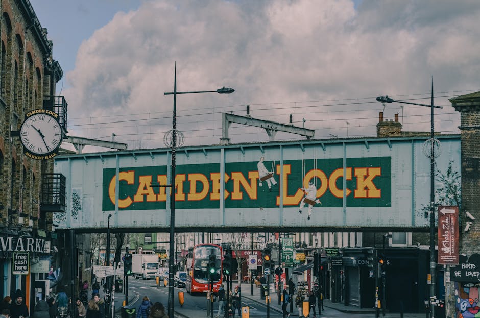 A busy urban street scene capturing the exterior of Camden Lock in Fulham, with a large painted Signage board reading 'CAMDEN LOCK' spanning above the street. In the foreground, a red double-decker bus is visible amidst pedestrians on the pavement, some carrying shopping bags. To the left, a street clock mounted on a building shows the time as approximately 2:15. The street is active with vehicles, including cars and additional buses, moving along the road. Overhead, streetlights and tram wires run across the scene, with cloudy skies above. The surroundings include brick buildings, shopfronts, and murals, indicating a lively commercial area suitable for home relocation and furniture transport services offered by Movers Fulham. The area is well-lit with natural daylight, capturing the typical atmosphere of a busy city access point for loading and unloading during house removals or furniture moving processes.