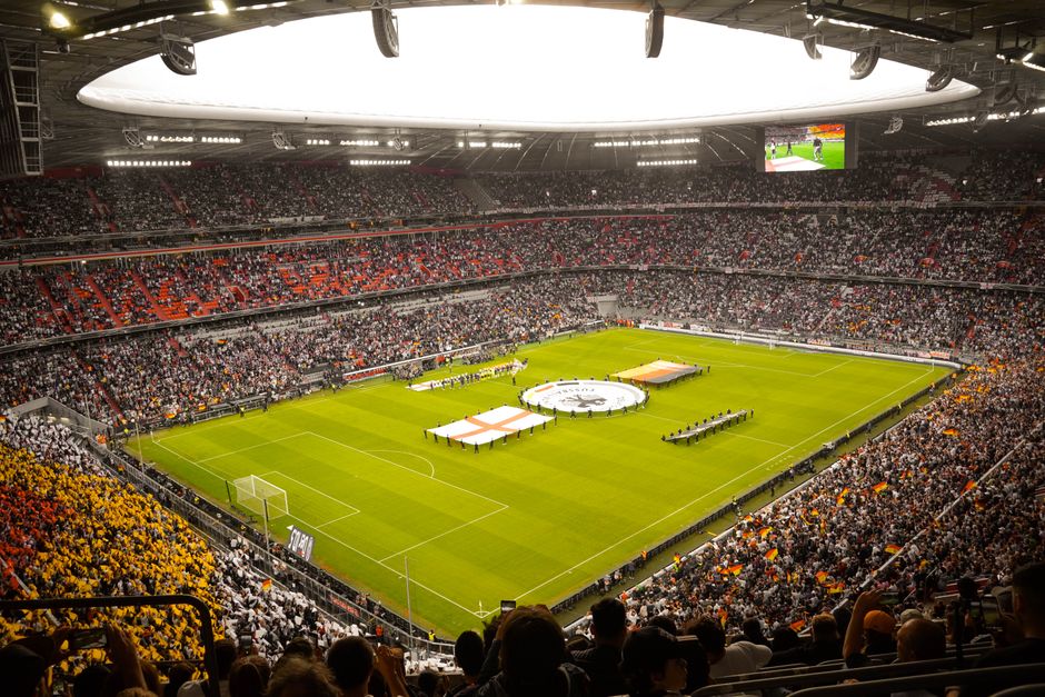 Inside a large football stadium during a match day, a well-maintained grass pitch is visible with white boundary lines and penalty areas. The stands surrounding the field are filled with spectators, many dressed in team colours, occupying seating sections that feature predominantly black, white, orange, and yellow seats. On the pitch, a circle of team banners and flags is arranged in the centre, with a few smaller flags placed along the sidelines. There are some people walking or standing near the banners, possibly preparing for a ceremonial or team presentation. Overhead, a large, bright, circular roof structure with a smooth, white surface provides partial cover, supported by a network of steel beams. Large stadium lights and electronic screens are visible, indicating a well-lit environment suitable for evening matches. The environment suggests a busy, organized atmosphere typical of a professional football event, with no visible furniture or equipment, and the focus remains on the playing field and the crowd. The image also subtly reflects the logistical aspects of event planning and crowd management associated with stadium operations.